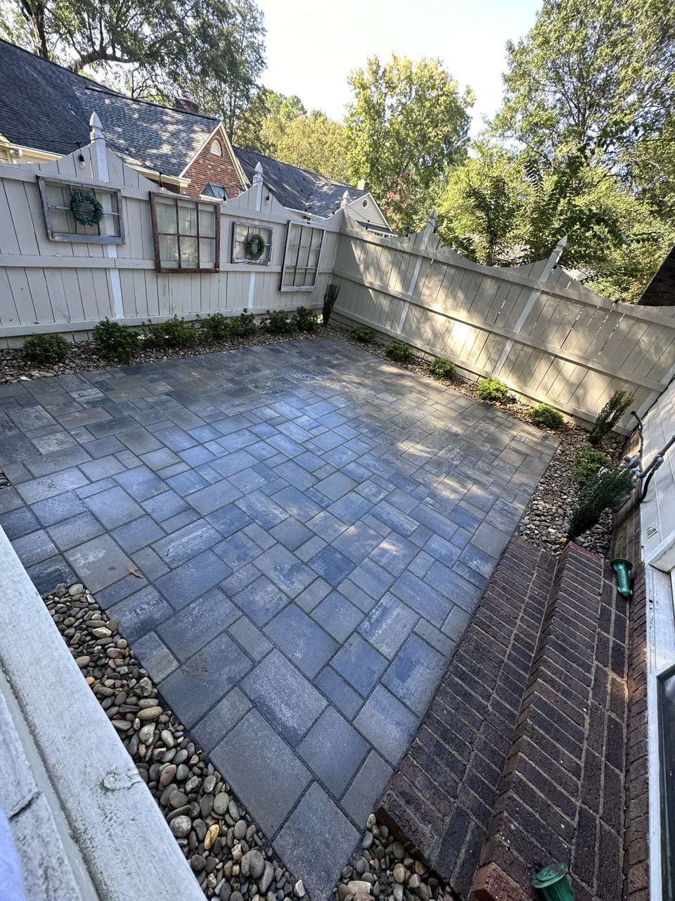 Paver courtyard framed by river rock, small shrubs and a white privacy fence
